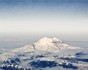 May include: A photograph of a snow-capped mountain peak rising above a sea of clouds. The mountain is surrounded by a vast expanse of white clouds, with a blue sky above.