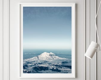 May include: A white armchair sits in front of a framed photograph of a snow-capped mountain peak rising above a sea of clouds. A small white side table with a book stack and a potted plant are in the foreground.