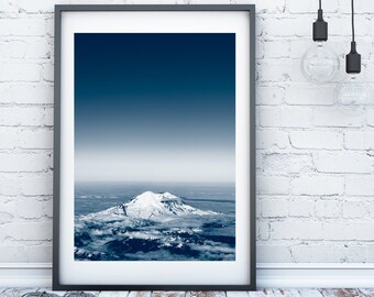 May include: A framed photograph of a snow-capped mountain peak rising above a sea of clouds. The sky is a deep blue, fading to a lighter blue at the horizon. The photo is in a black frame and is hanging on a white brick wall.