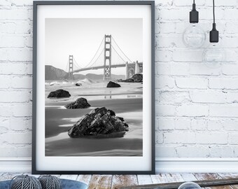 May include: Black and white framed print of the Golden Gate Bridge in San Francisco. The photograph captures the bridge against a backdrop of the ocean and beach, with rocks in the foreground. The frame is dark gray.