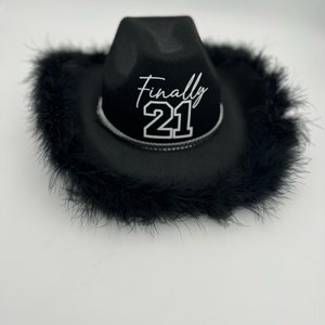 May include: Black cowboy hat with a black feather trim. The hat has the text "Finally 21" in white lettering.