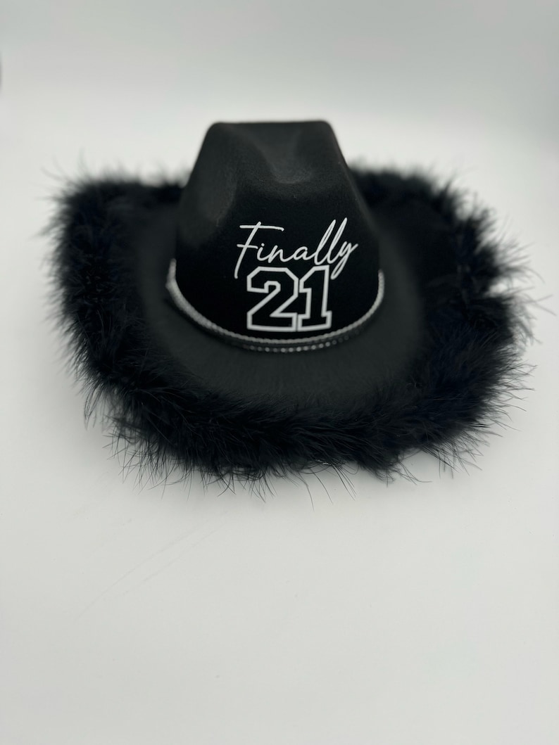 May include: Black cowboy hat with a black feather trim. The hat has the text "Finally 21" in white lettering.