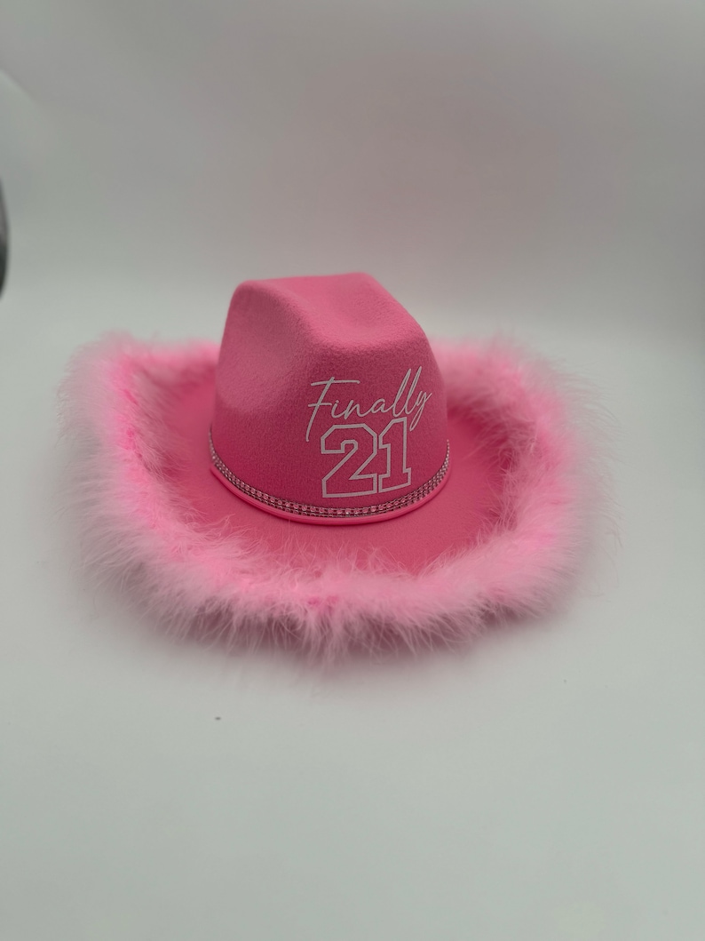 May include: Pink cowboy hat with a white feather trim and the text "Finally 21" embroidered on the front.