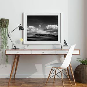 May include: A black and white photograph of a mountain range under a cloudy sky, framed in a white frame and hanging on a white wall above a white desk with a black lamp and a white chair.