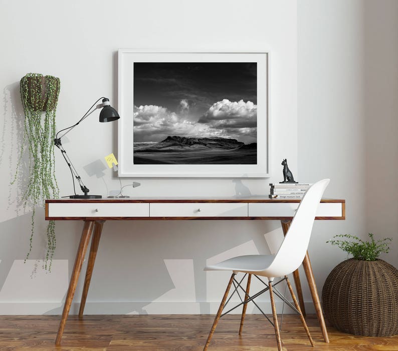 May include: A black and white photograph of a mountain range under a cloudy sky, framed in a white frame and hanging on a white wall above a white desk with a black lamp and a white chair.