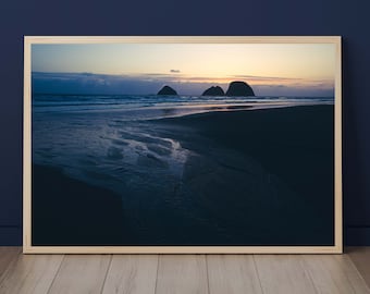 May include: A framed photograph of a beach scene at sunset. The photo shows a dark, sandy beach with a small stream of water flowing towards the ocean. In the distance, three rocky islands are silhouetted against the orange and pink sky.