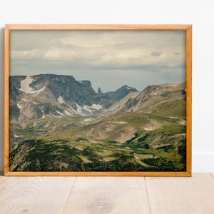 May include: A framed photograph of a mountain range with a cloudy sky. The mountains are covered in green vegetation and snow. The photo is in a wooden frame.