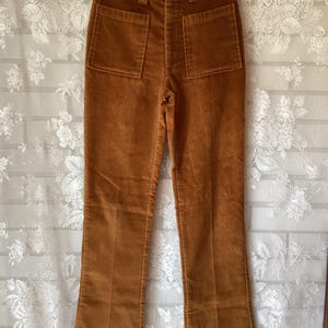 May include: A pair of brown corduroy pants with two back pockets and a classic fit. The pants are hanging on a hanger against a white floral background. The corduroy fabric has a soft, textured appearance.