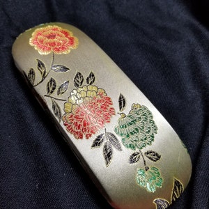 May include: A rectangular, light-colored eyeglasses case with a floral design. The case features embroidered flowers in red, green, and gold, with black and gold leaves. The case is closed and rests on a dark fabric surface.