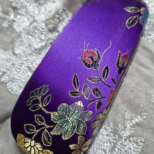 May include: A purple, rectangular glasses case with floral embroidery. The case is a deep violet color and features embroidered flowers in shades of green, gold, red, and blue. The case has a satin-like texture.