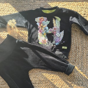 May include: Black velour baby outfit with a long-sleeved sweatshirt featuring a floral letter H design. The outfit includes matching pants.