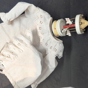 以下が含まれることがあります： アイレットレースのトリムが付いた白いコットン製のベビーワンピースと、それに合わせたボンネット。ワンピースは長袖で、ギャザーの入ったネックライン。ボンネットはレースのトリムと後ろにリボンが付いています。背景には、ペイントされた馬がついた木製のカルーセルがあります。