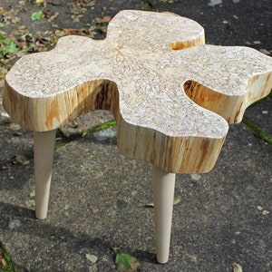 May include: A unique, natural wood table with a live edge top and three legs. The table top has a light brown color with a natural wood grain pattern. The legs are made of a lighter wood and are tapered.
