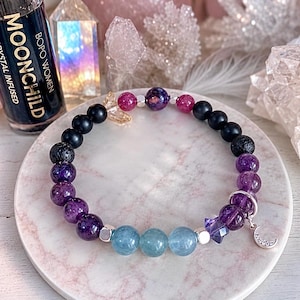 May include: A beaded bracelet featuring a mix of black, purple, light blue, and pink spherical beads, including textured lava stone beads. The bracelet is accented with silver-toned spacers, a small star-shaped bead, and a silver crescent moon charm.