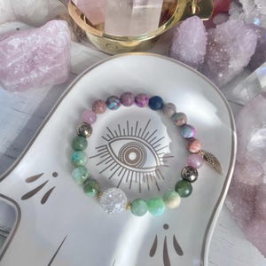 Puede incluir: Una pulsera de cuentas con una mezcla de piedras redondas en tonos verde, rosa, morado y azul, que incluye una cuenta de cristal craquelado transparente y un dije de ala en metal dorado. La pulsera se presenta sobre una bandeja de cerámica blanca.