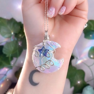 May include: A crescent moon-shaped pendant made of iridescent, translucent material wrapped in silver-coloured wire. A small, dark blue star-shaped charm is attached to the front. The pendant hangs from a thin silver-coloured metal chain.