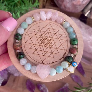 May include: A gemstone bracelet with pink, green, and blue beads, including a heart-shaped pink stone and a gold evil eye charm. The bracelet is displayed on a wooden coaster. The coaster has a geometric design and the text "the weekend mystic".