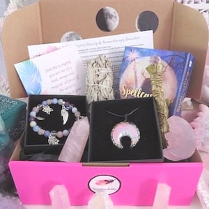 Puede incluir: Una caja de regalo rosa llena de varios artículos, incluyendo un brazalete de cristal, un collar con un colgante de luna creciente, un palo de salvia y un libro titulado "Spellcraft". La caja está rodeada de cristales y otros artículos espirituales.