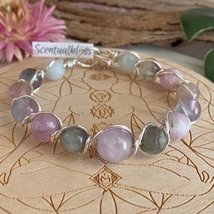 Peut inclure: Un bracelet fait à la main avec des pierres précieuses colorées, notamment des perles roses, violettes et grises. Les perles sont enfilées avec du fil d'argent. Le bracelet est présenté sur une surface en bois avec des motifs gravés. Le texte "Scentualbliss" est visible.
