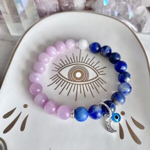 May include: A beaded bracelet featuring a mix of lavender, white, and blue gemstones. The bracelet includes a silver charm with a crescent moon and an evil eye. The bracelet is displayed on a white dish with an eye design.