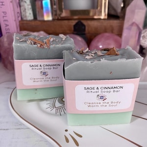 May include: Two sage and cinnamon ritual soap bars. The top layer is a muted green colour, with bits of cinnamon bark. The bottom layer is a lighter green. Each bar has a pink label with text that reads "Sage & Cinnamon Ritual Soap Bar".
