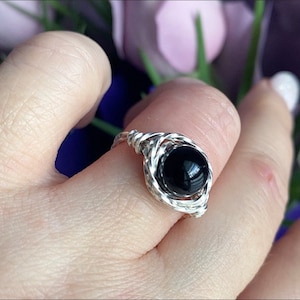 May include: A wire-wrapped ring featuring a single, smooth, round black stone at its centre. The band and the setting are constructed from a metallic silver-toned wire that is twisted and coiled around the stone to hold it securely in place.