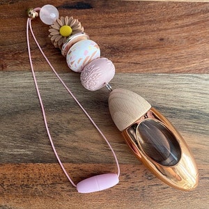 May include: A pink and gold car air freshener with a wooden top and a pink beaded chain. The beads feature floral and abstract patterns.