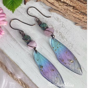 May include: Handmade earrings with iridescent wing-shaped pendants in blue, purple, and gold. Copper-coloured hooks support pink and green gemstone beads. The earrings are finished with the text "Scentualbliss".