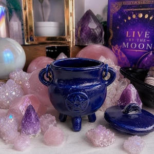 May include: A deep blue ceramic cauldron with a raised pentacle symbol on the front, featuring three small legs and two side handles. The cauldron includes a matching lid and is surrounded by various raw crystals, including rose quartz and amethyst.