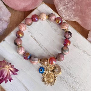 May include: A beaded bracelet featuring pink and iridescent beads. The bracelet has gold accents and charms, including a sunflower, a heart, and an evil eye. The bracelet is displayed on a white wooden surface.