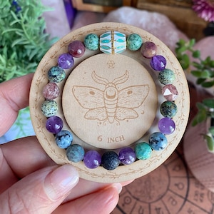 May include: A beaded bracelet featuring a mix of round purple, green, blue, and speckled stones, along with a central white and gold-toned metal bead. The bracelet is displayed on a circular wooden stand marked 6 INCH with a moth illustration.