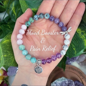 May include: A beaded bracelet with a variety of colorful stones, including pink, turquoise, and purple, is displayed. A silver dragonfly charm and a lotus flower charm are attached. The text "Mood Booster & Pain Relief" is superimposed on the image.
