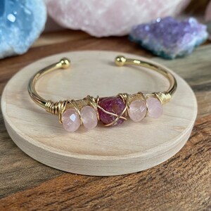 May include: Gold wire wrapped bracelet with pink and red gemstone beads. The bracelet is on a wooden surface.