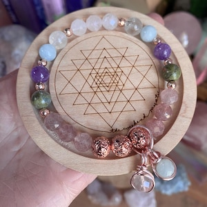 May include: A multi-gemstone bracelet featuring amethyst, rose quartz, and aquamarine beads, presented on a wooden coaster with a geometric pattern. The bracelet includes rose gold elements and a charm with two hanging stones.