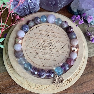 Peut inclure: Un bracelet de perles composé de pierres précieuses violettes, roses et transparentes. Il repose sur un présentoir en bois avec un motif géométrique et un charm lotus argenté. Le présentoir porte l'inscription "the weekend mystic".