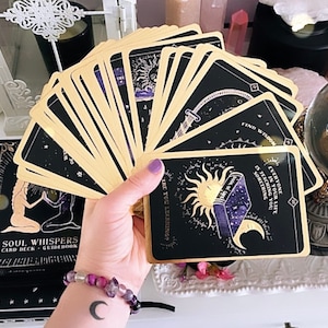May include: A deck of Soul Whispers tarot cards fanned out, featuring black card faces with gold accents and celestial illustrations. The guidebook is visible in the background. The cards have text that reads "Find Wisdom" and "Everyone in your life is teaching you something."