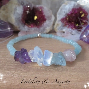 May include: A light blue beaded bracelet with a silver clasp. The bracelet features a variety of crystals, including amethyst, moonstone, aquamarine, and rose quartz. The text "Fertility & Anxiety" is written below the bracelet.