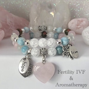 May include: Two bracelets featuring clear crystal beads, blue and white beads, and silver charms. The charms include a heart-shaped pink stone, a butterfly, and two oval charms that say "hope" and "believe". The bracelets are displayed on a white surface with pink and white stones in the background. The text "Fertility IVF & Aromatherapy" is below the bracelets.
