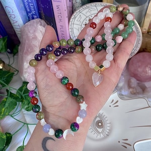 May include: Several beaded bracelets in shades of pink, green, red, purple, and white. One bracelet has a heart-shaped charm. The bracelets are shown on an open hand.