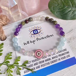May include: A multi-gemstone bracelet featuring purple, blue, and clear beads. The bracelet is presented with a card that reads "Evil Eye Protection". The bracelet includes a pink charm with an eye design. The card's text promotes good luck.