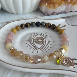 May include: A beaded bracelet featuring a mix of round tiger eye, lava, and faceted quartz beads in shades of brown, tan, pink, and yellow. The bracelet includes a gold-toned metal charm with a sun design and a small green crystal accent.