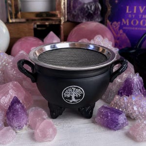 May include: A matte black cauldron with two side handles and three feet, featuring a silver tree of life emblem on the front and a circular silver-rimmed metal mesh insert covering the top opening, surrounded by various raw rose quartz crystals.