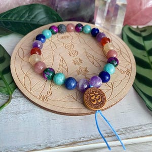 May include: A multi-coloured gemstone bracelet sits on a wooden coaster. The bracelet features beads in shades of purple, blue, green, and pink. The coaster has an engraved illustration of a human figure with chakra symbols. A wooden charm with the Om symbol is attached.
