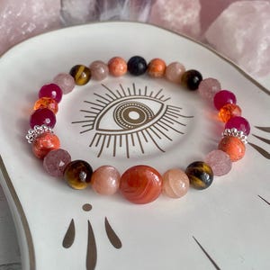 May include: A beaded bracelet featuring a variety of round gemstones in shades of orange, red, brown, and pink. The bracelet includes faceted beads, smooth polished stones, and two silver-toned metal spacers with a textured, beaded edge design.