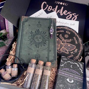 Pode incluir: Uma caixa de presente contendo um diário verde com um motivo de sol, um tabuleiro circular preto com um gráfico de mariposa, um baralho de cartas oráculo Cosmic Guidance, três frascos de vidro com ervas secas e um caldeirão preto.