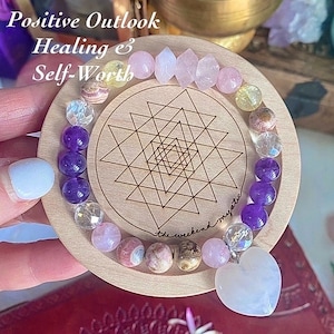 May include: A beaded bracelet featuring various gemstones in shades of purple, pink, yellow, and clear, with a heart-shaped pendant. The bracelet rests on a circular wooden disc engraved with a geometric star pattern and the text Positive Outlook Healing & Self-Worth.