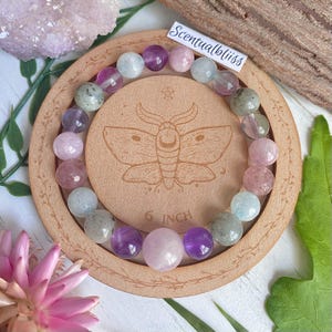 May include: A gemstone bracelet with pink, purple, and blue beads rests on a wooden coaster. The coaster has a moth design and reads "6 INCH" (approx. 15 cm). The brand name "Scentualbliss" is visible. The bracelet is a piece of jewelry.