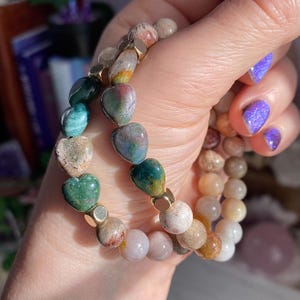 May include: Two beaded bracelets. One bracelet features heart-shaped stones in shades of green, tan, and pink, separated by gold-colored beads. The other bracelet has round, light-colored beads. The bracelets are held in a hand with purple nail polish.
