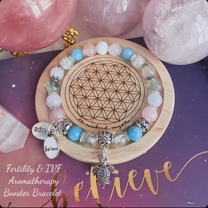 May include: A light blue, pink and green crystal bracelet with silver charms and a turtle pendant. The bracelet is on a wooden coaster with a flower of life design. The text "Believe" is on a silver charm. The text "Fertility & IVF Aromatherapy Booster Bracelet" is below the coaster.
