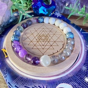 May include: A colourful gemstone bracelet with purple, white, and blue beads. The bracelet is displayed on a wooden coaster with a geometric design and the text "weekend mystic".